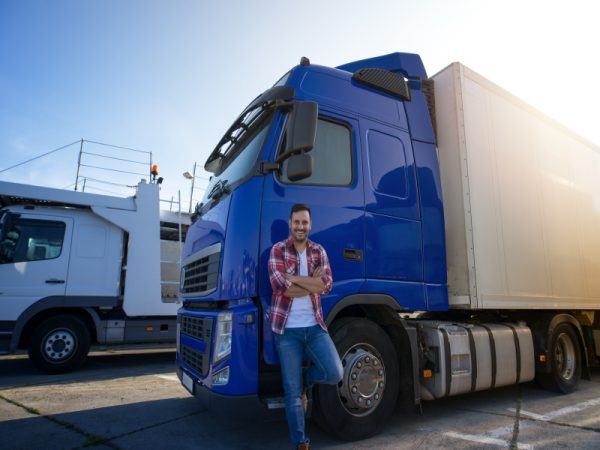 Truck driver with crossed arms and smile on his face standing by his truck on a break ready for a ride. Transportation services.